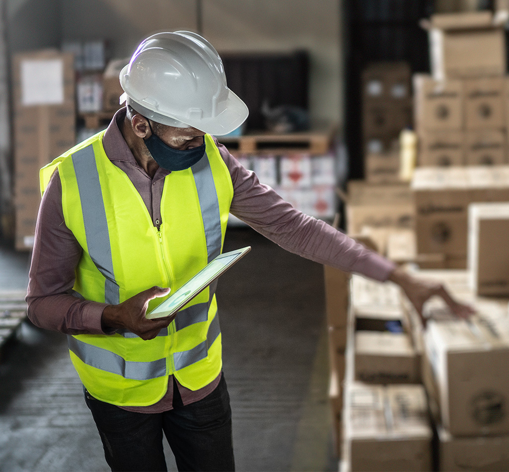 Warehouse worker wearing face mask and protective workwear checking products using digital tablet
